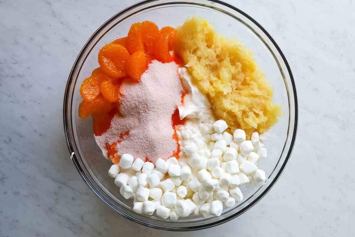 All of the ingredients for an orange fluff salad together inside a glass bowl on a counter.