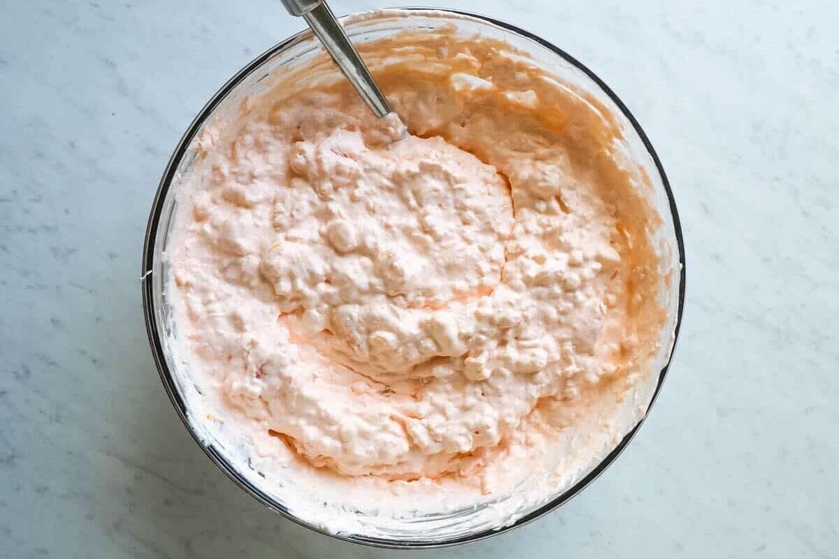 Freshly mixed orange jello salad inside a glass bowl with a silver mixing spoon.