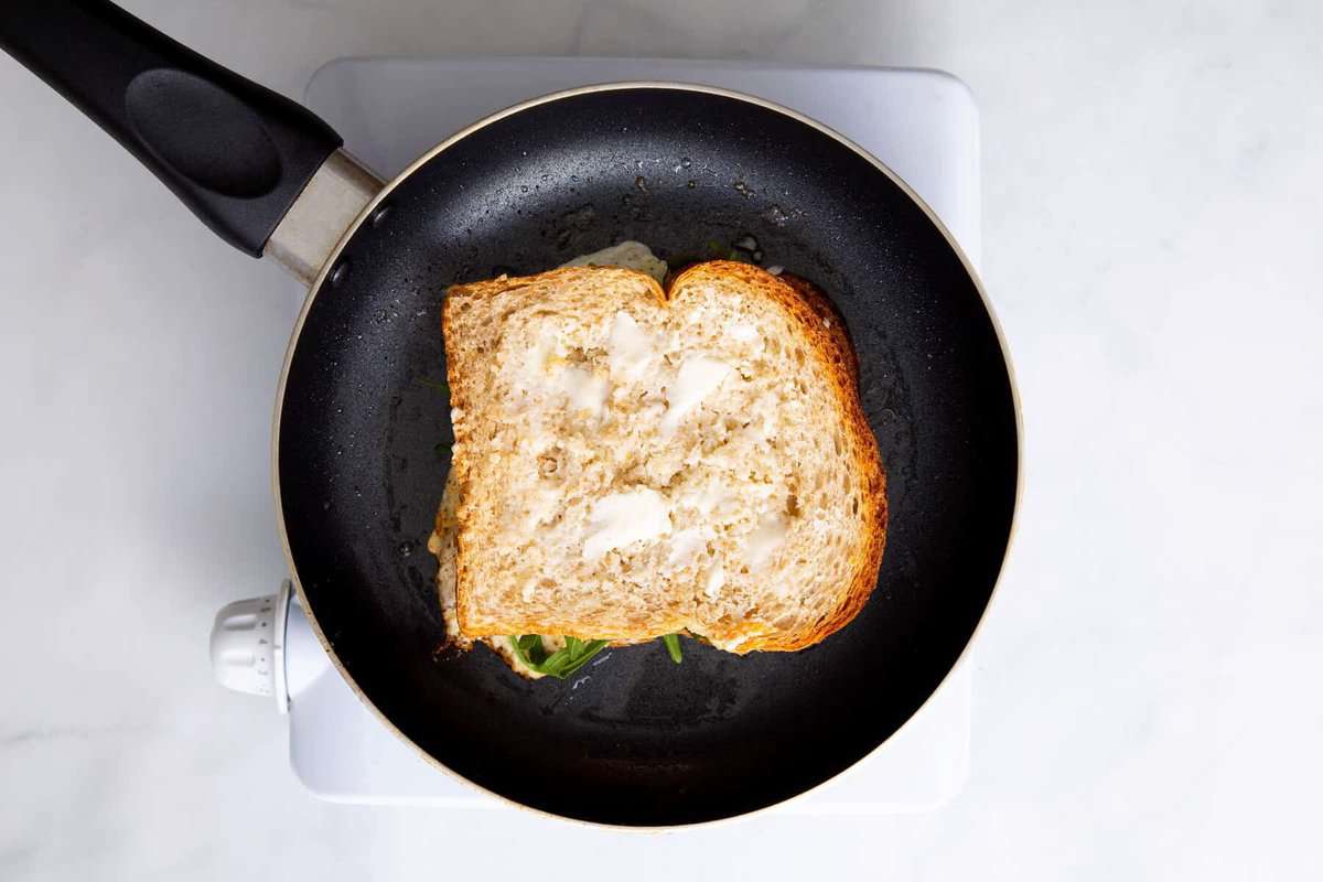 Buttered bread added on top of egg and cheese sandwich in pan.