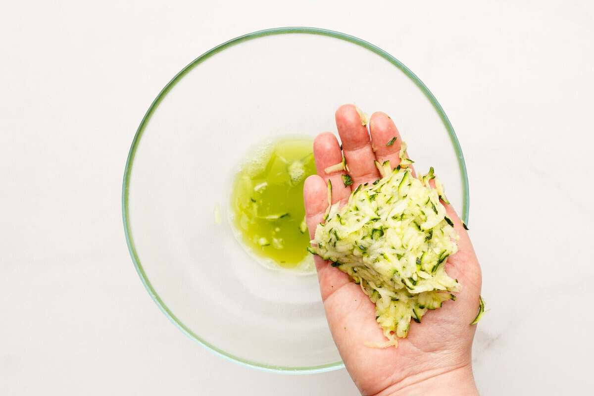 Hand holding shredded zucchini over bowl with liquid.