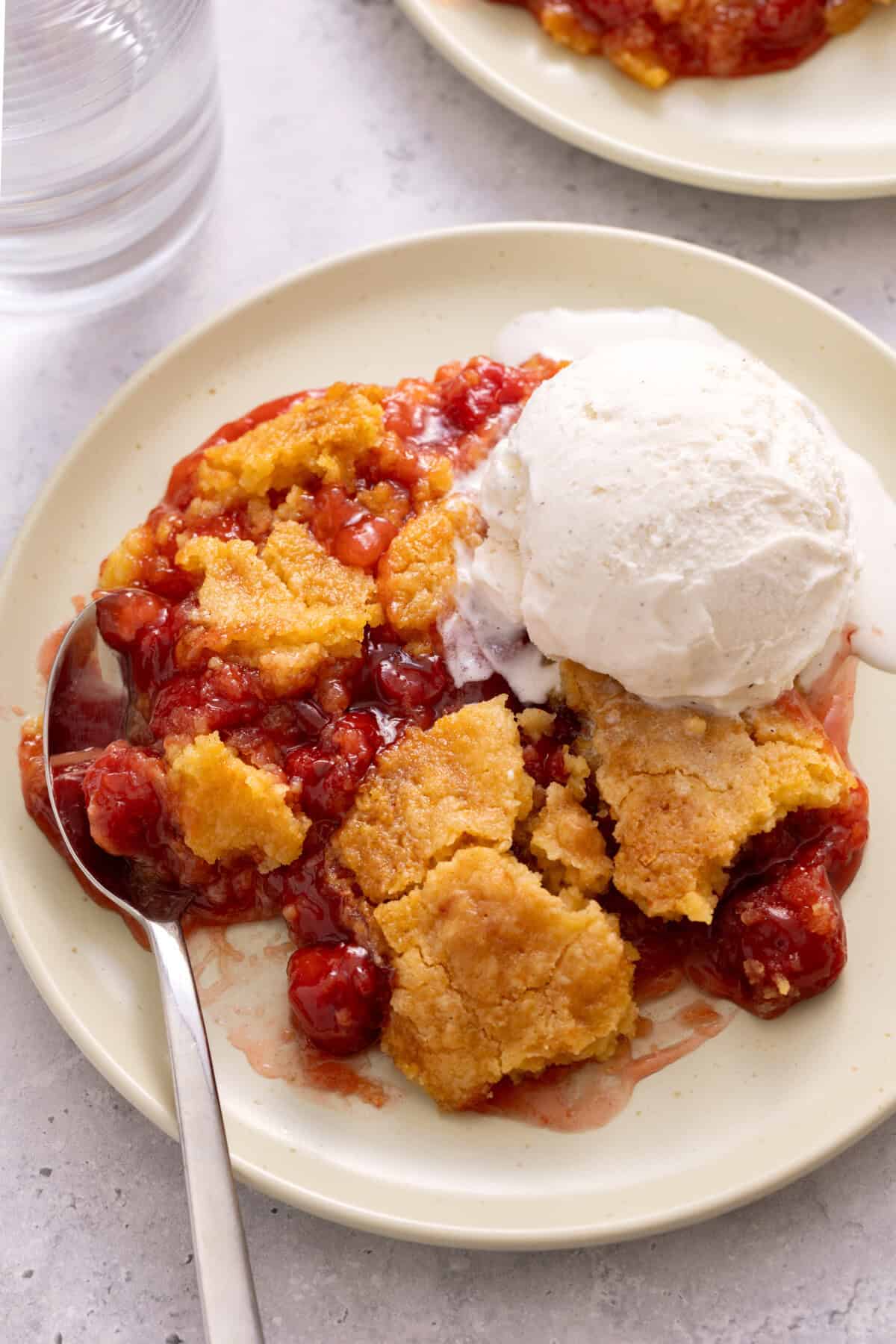 Serving of cherry dump cake in bowl with scoop of vanilla ice cream and spoon.
