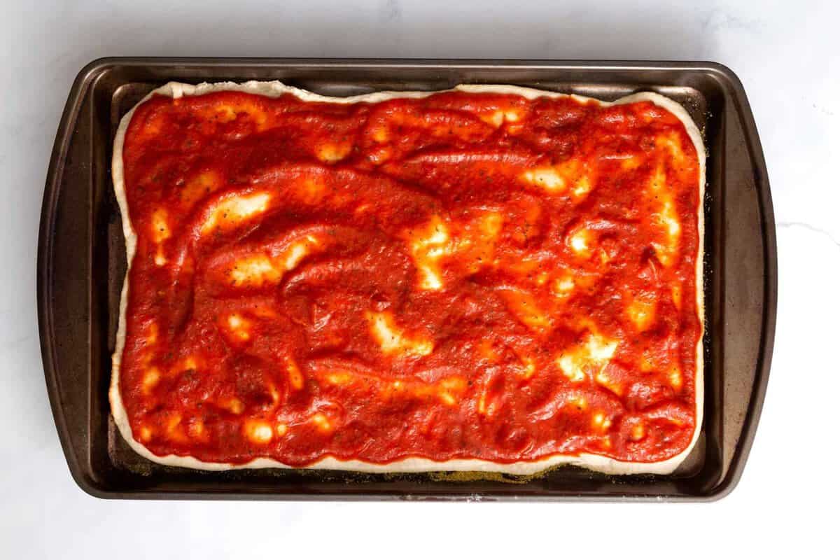 Pizza dough stretched in sheet pan and topped with tomato sauce.