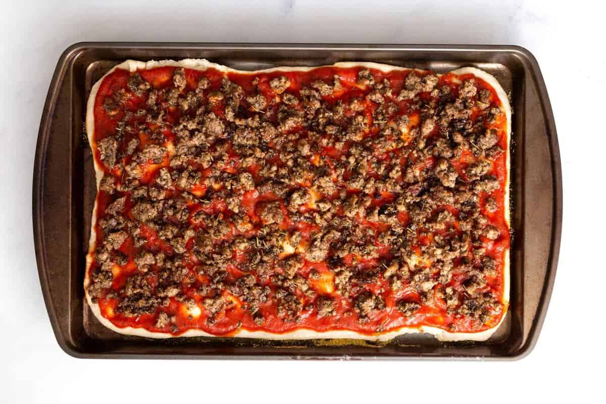 Pizza dough stretched in sheet pan and topped with tomato sauce and sausage.