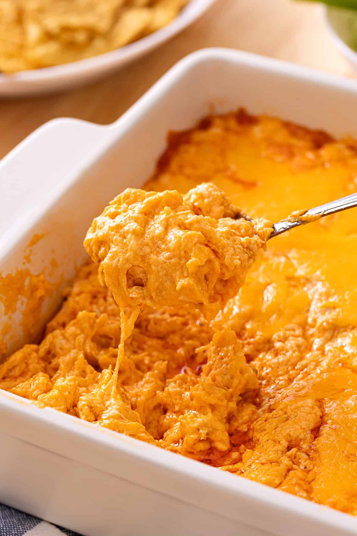 A spoon lifting cheesy buffalo chicken dip from a white baking dish.